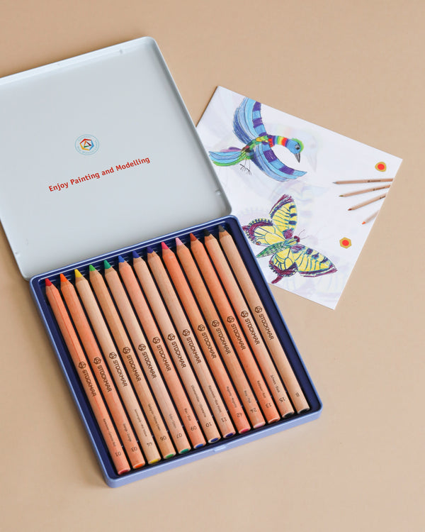 A set of Stockmar Triangular Coloured Pencils - 12, made from sustainably managed forests, neatly arranged in a metal box, next to a hand-drawn picture of a colorful bird and butterflies on a piece of paper