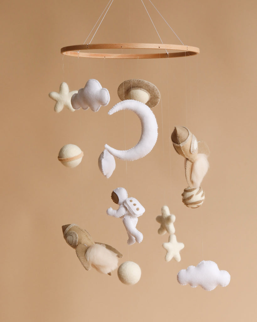 A nursery mobile featuring plush toys including stars, a moon, clouds, and an astronaut, all suspended from a circular wooden frame against a soft beige background.