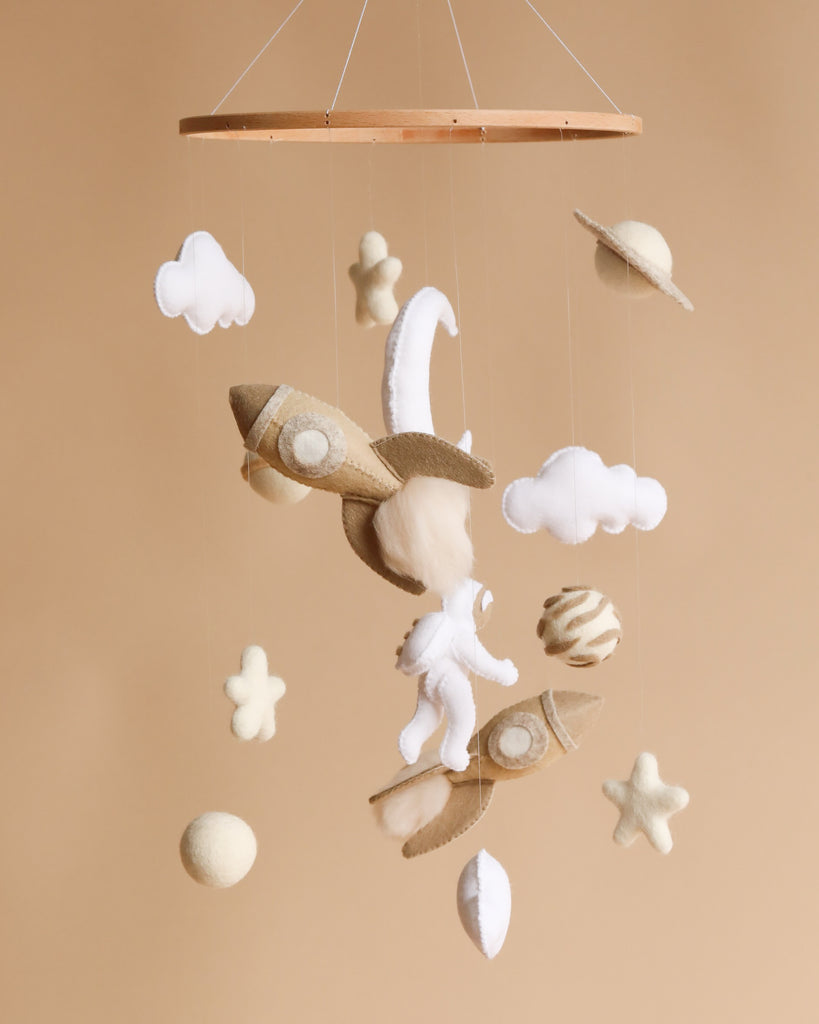 A baby mobile featuring plush airplane, clouds, and celestial bodies, with a felt astronaut dangling amid the elements, set against a soft beige background. This Handmade Mobile - Among the Stars - Final Sale is handmade in Europe.