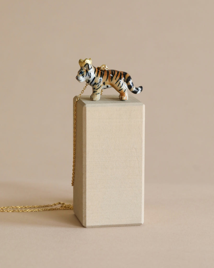A ceramic Tiger King Necklace atop a canvas-textured rectangular box with a 24k gold plated chain draped beside it, set against a soft beige background.