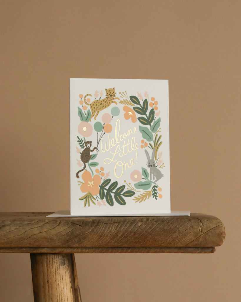 The Menagerie Baby Card reads "Welcome Little One!" and features illustrated animals, leaves, and flowers in pastel colors with gold foil accents. It stands on a rustic wooden stool against a beige background. The interior is blank.