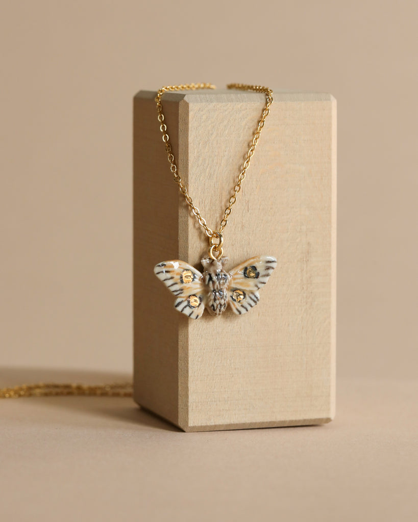 A Solar Moth Necklace with detailed wings hangs from a beige jewelry box against a soft beige background, emphasizing its elegant design.