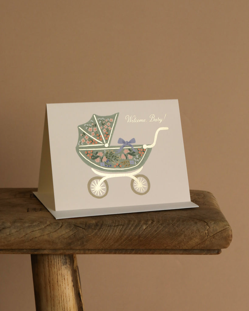 The Floral Pram Card features an illustrated baby stroller with floral patterns, a bow, gold foil accents, and "Welcome Baby!" on the front. It comes with a soft white envelope and is perfect for celebrating new arrivals.