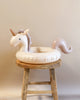 An Inflatable Junior Swim Ring - Unicorn sits atop a rustic wooden stool against a plain beige background. The unicorn has a white body with a soft pink and gray mane and tail.