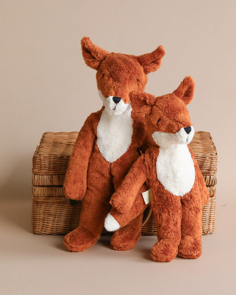 Two Senger Naturwelt Stuffed Animal - Foxes sitting snugly next to each other on a wicker basket, against a neutral beige background. The foxes are in a sitting position and appear soft and cuddly.