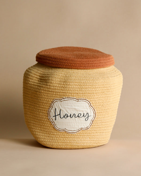 A textured Basket Honey Pot basket with a beige body and a brown cloth lid. The front features a white label with "honey" written in cursive script. The basket is set against a neutral background