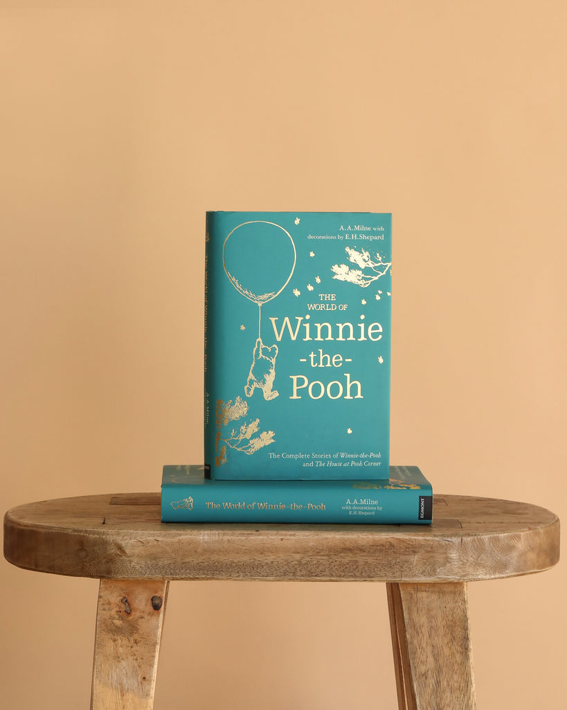 A hardcover edition of The World of Winnie-the-Pooh stands upright on a wooden stool, with another copy lying flat below it, against a plain beige background.
