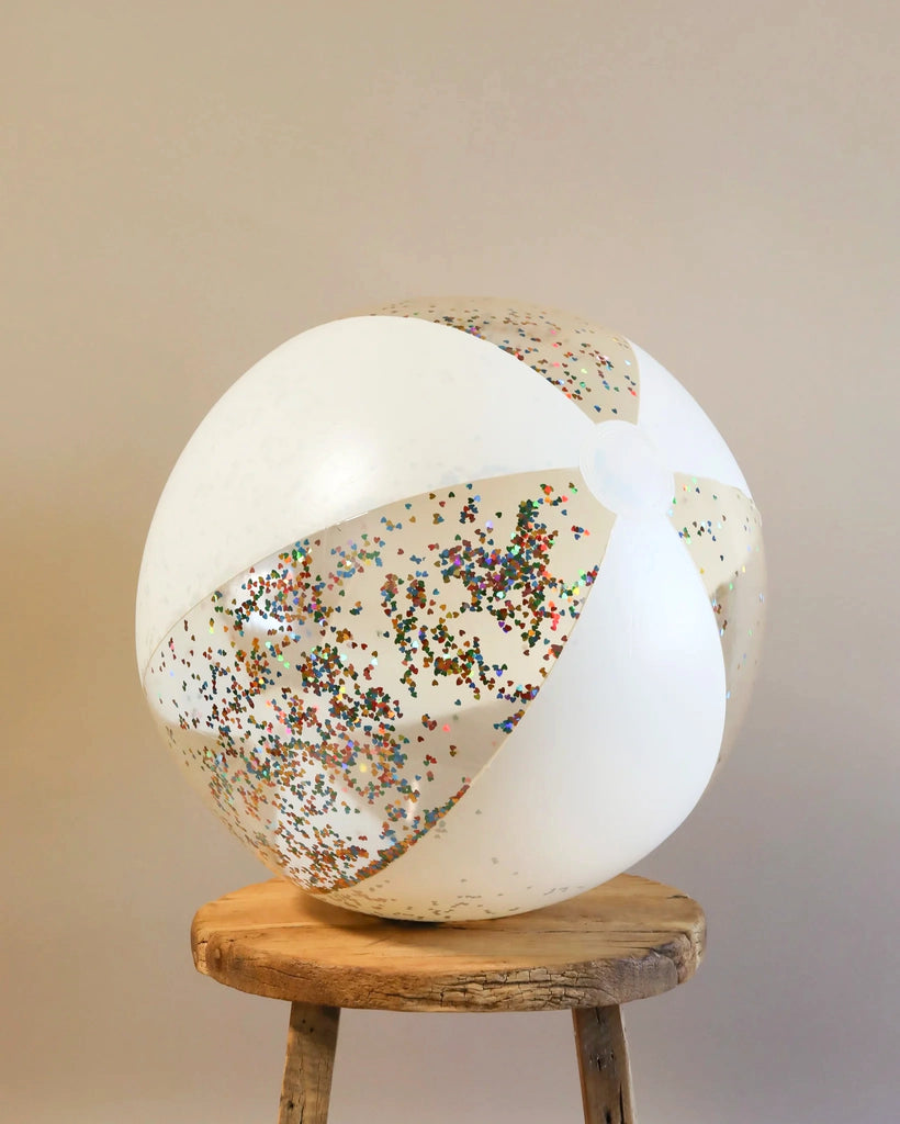 A large, durable white Inflatable Large Beach Ball with Transparent Confetti Hearts scattered over it, sitting on a rustic wooden stool against a neutral background.