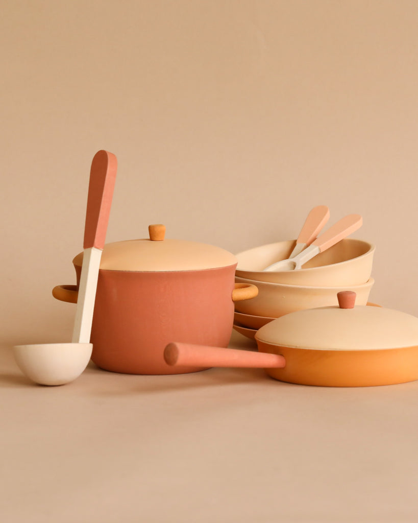 A Handmade Wooden Kitchen Essentials - Flower set in muted earth tones, including a large pot with a lid, a frying pan, a ladle, and bowls with spoons, all arranged neatly against a beige background. The items have a minimalist design and complementary colors in shades of beige and terracotta.