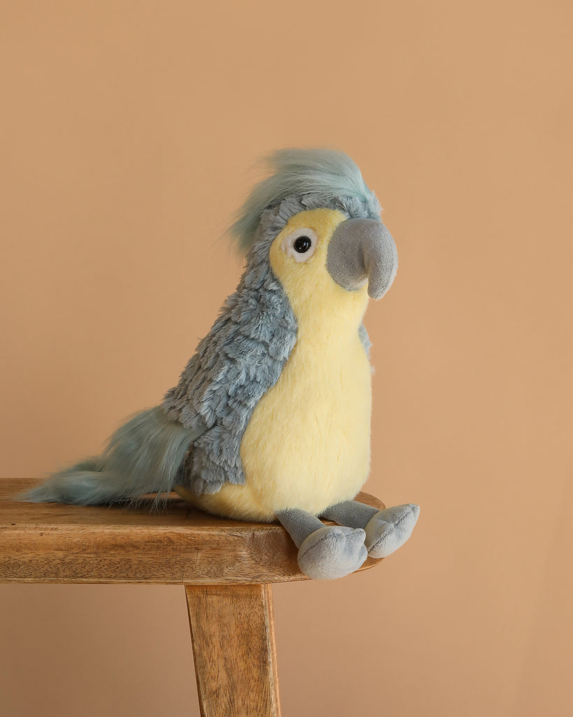 The Blu Cockatoo Bird Plush Toy, featuring pale yellow and light blue fur, sits on the edge of a wooden table with a plain beige background.