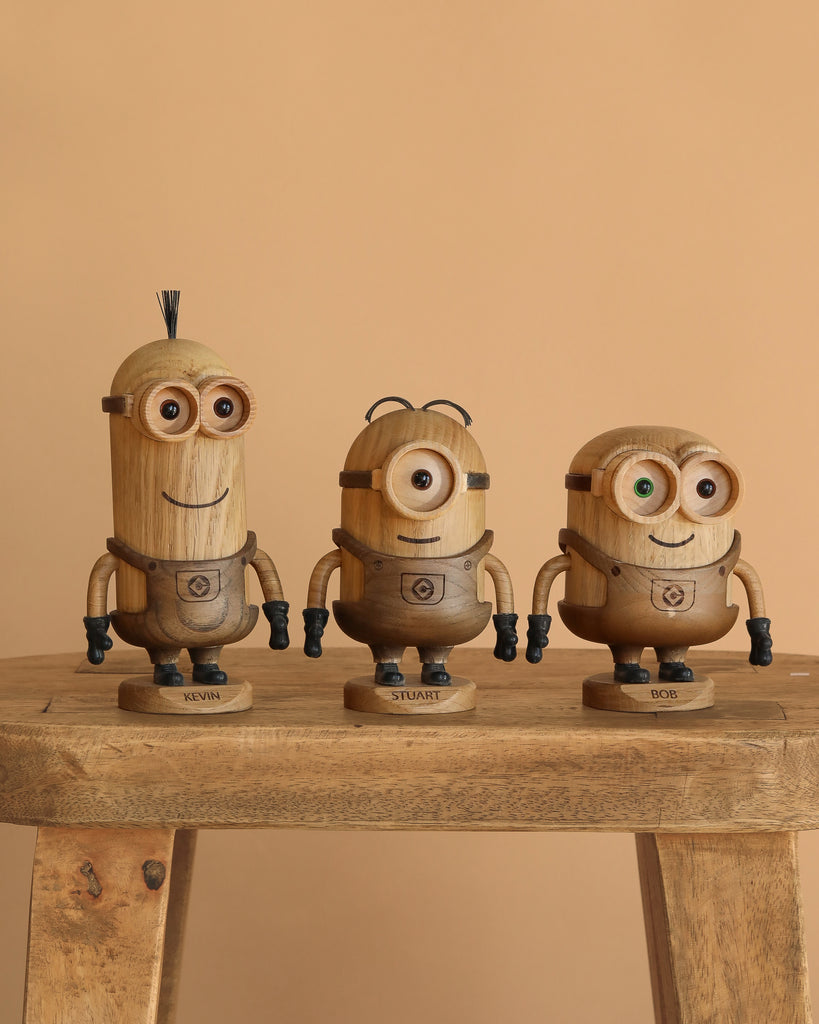 The Boyhood Minions Stuart, Small—crafted from European FSC White Oak—appears on a wooden bench with nameplates for Kevin, Stuart, and Bob, resembling Minions from Despicable Me against a plain beige background.