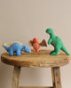Three Olli Ella Holdie Pre-Historic Animals—a blue stegosaurus, a pink triceratops, and a green T-Rex—standing on a rustic wooden table.