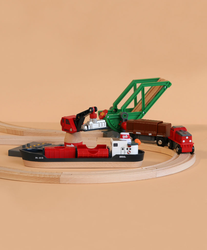 BRIO World Train Set Cargo Harbour Set: Wooden toy train set featuring tracks, a red and black cargo ship, red crane, green bridge, brown freight cars, and a red engine on a light beige background.