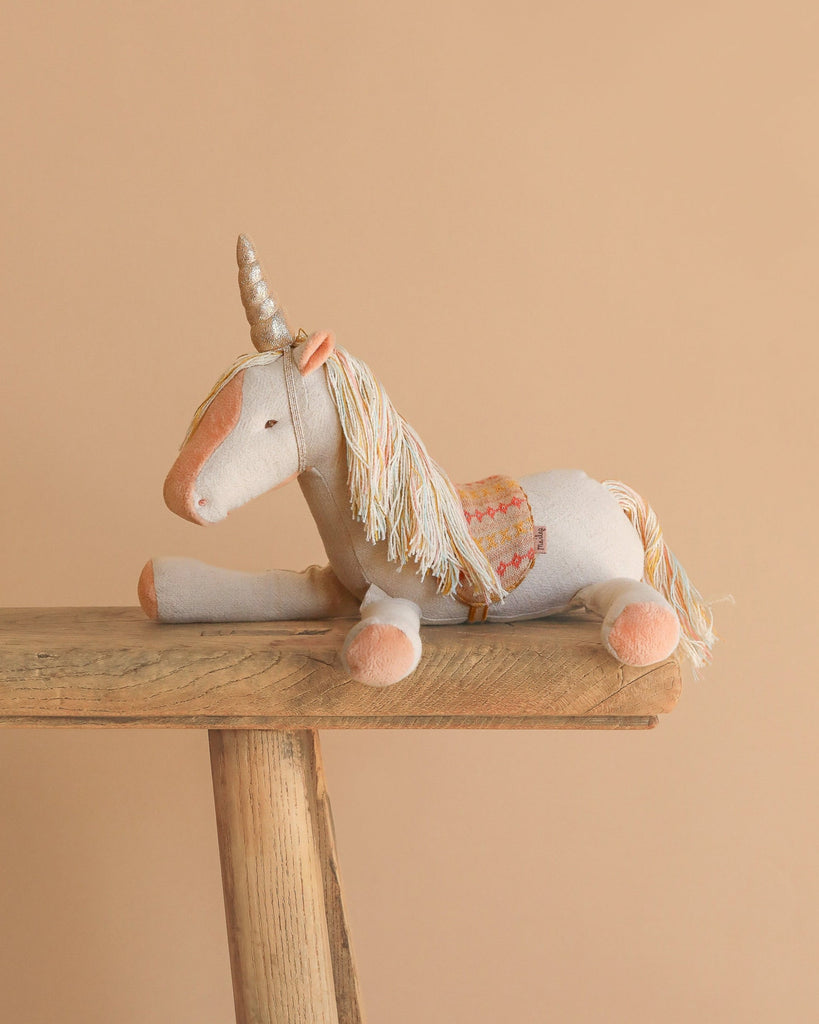 The Maileg Unicorn, Medium—a plush toy with a golden horn, pastel mane and tail, and patterned saddle—rests on a wooden bench against a plain beige background, inviting magical stories.