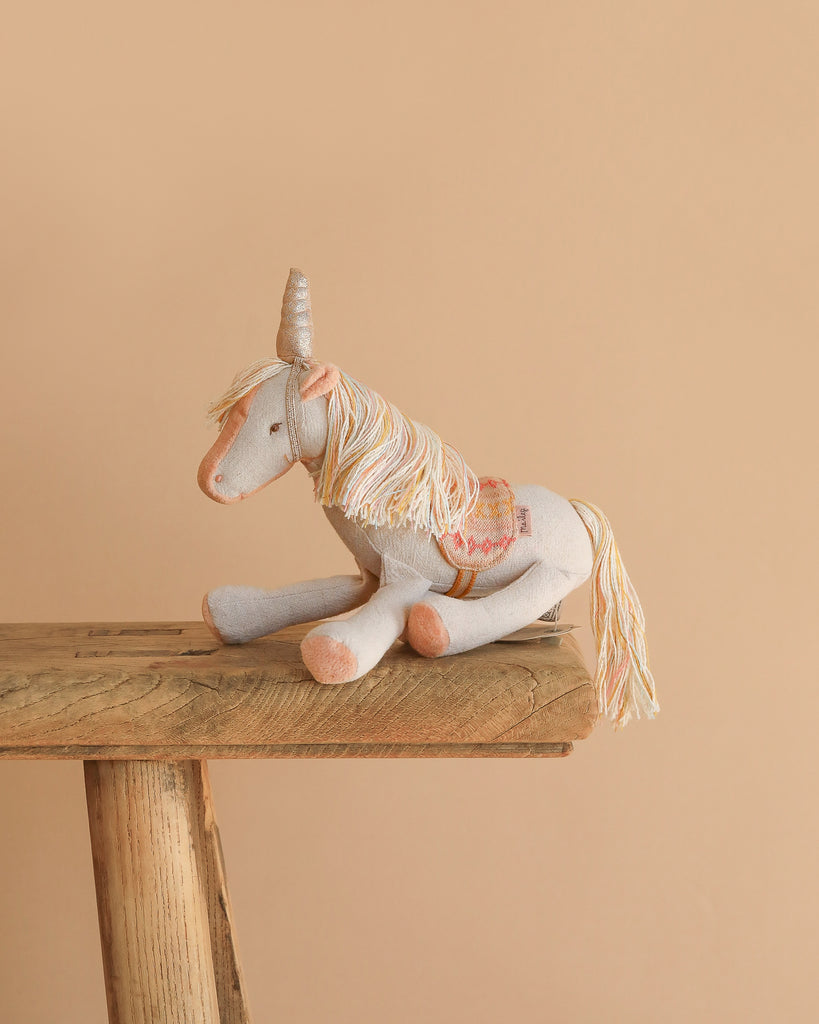The Maileg Unicorn, Small—a soft toy with a gray body, cream mane and tail, golden horn, and decorative saddle—sits on a rustic wooden stool against a beige background.