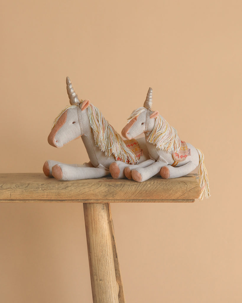 Two Maileg Unicorn, Small plush toys with cream bodies, golden horns, and silver accents sit side by side on a rustic wooden bench against a peach-colored background.