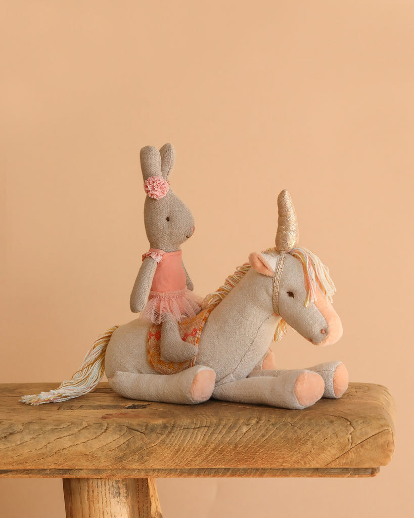A Maileg Unicorn, Small with a golden horn serves as a seat for a stuffed bunny in a pink dress and headband, both displayed on rustic wood against a beige backdrop. Soft, colorful hues enhance the scene’s whimsical charm.