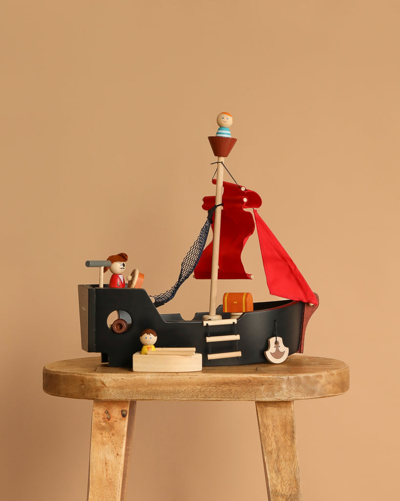 The Pirate Ship Set, featuring red sails and small figurines, sits on a rustic wooden stool against a plain beige background, making it an enchanting toy for children.