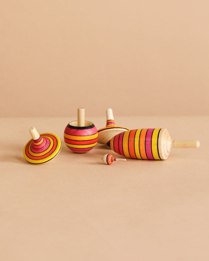 Five vibrant wooden tops from the Mader Spinning Top Champion Learning Set - Fire are displayed on a beige surface. Each top features bold red, orange, yellow, black, and pink stripes, ideal for all skill levels.
