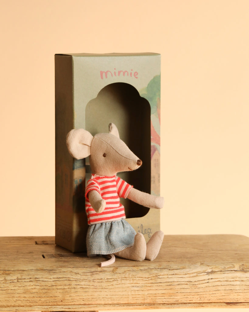 A Maileg Sister’s Day Out Mouse, dressed in a red-and-white striped shirt and blue skirt, sits before a green "mimie" box on a wooden surface with a beige background. Ideal for Big Sister Mouse collectors.