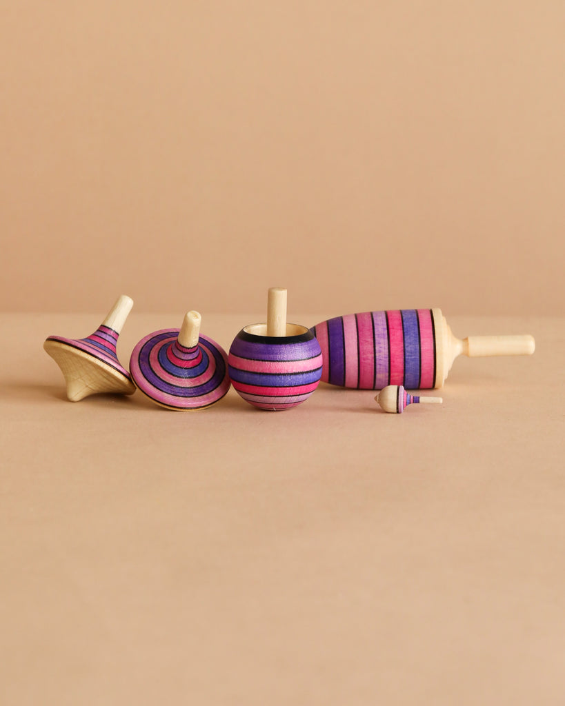 The Mader Spinning Top Champion Learning Set - Lilac features four fun tops in purple, pink, and blue stripes, including one tiny top. All meet toy safety standards and add playful color to any collection on a plain beige surface.