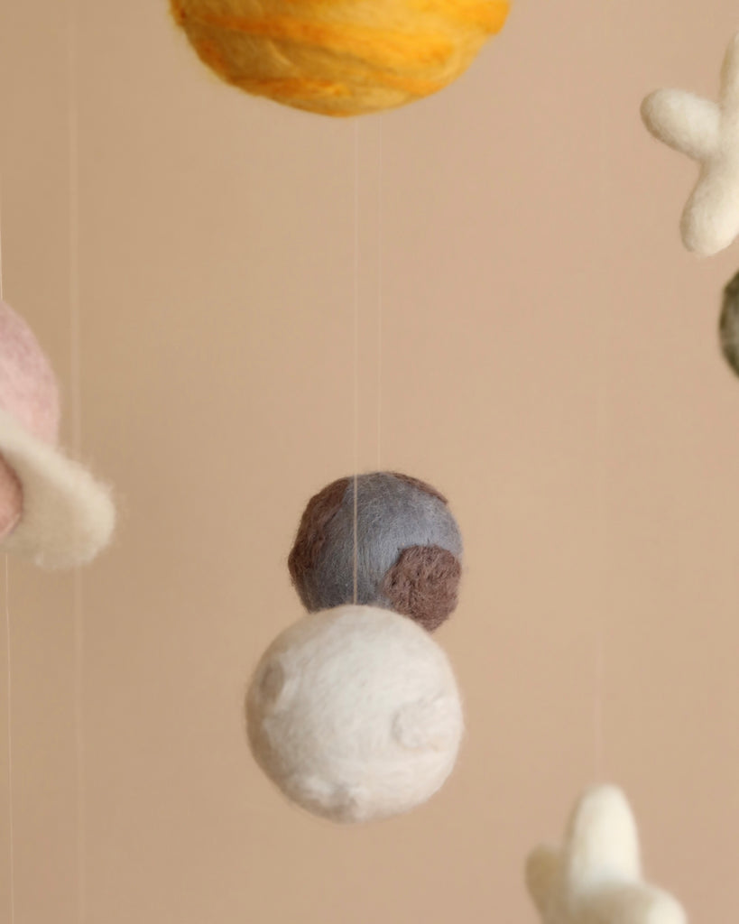 Close-up image of a Handmade Mobile - Outer Space - Final Sale with felted wool planets in soft pastel colors suspended on thin strings against a light beige background.
