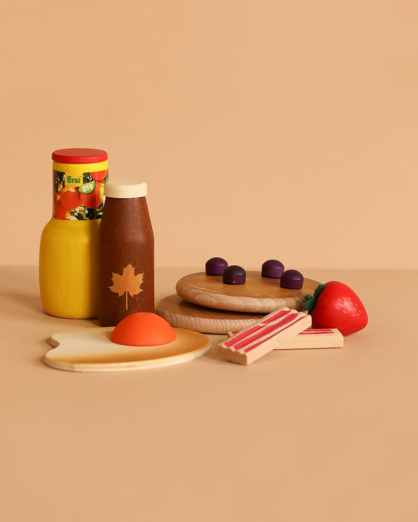 The Erzi American Breakfast Set features wooden toy foods like a fried egg, bacon strips, strawberry, maple syrup bottle, yellow jar, and purple pieces on plates, all arranged against a beige background.