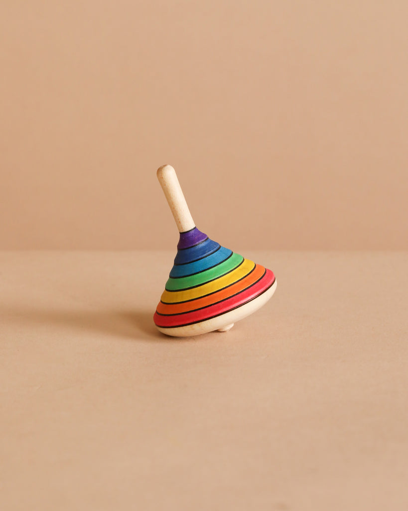 The Spinning Top - Rainbow, crafted from durable maple wood with vibrant rainbow stripes, rests on a light tan surface against a matching background.