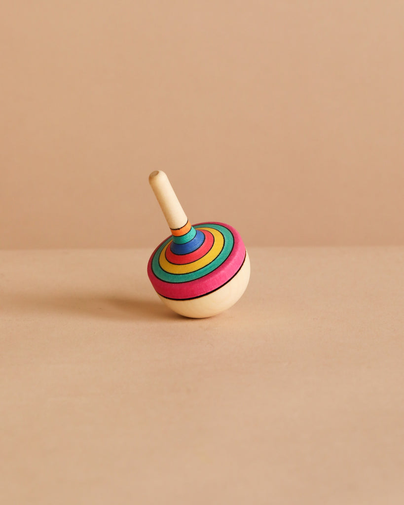 The Traditional Spinning Top – Striped features colorful concentric rings in red, yellow, green, and blue on crafted maple wood. It meets strict toy safety standards and is shown on a beige surface with a matching background.