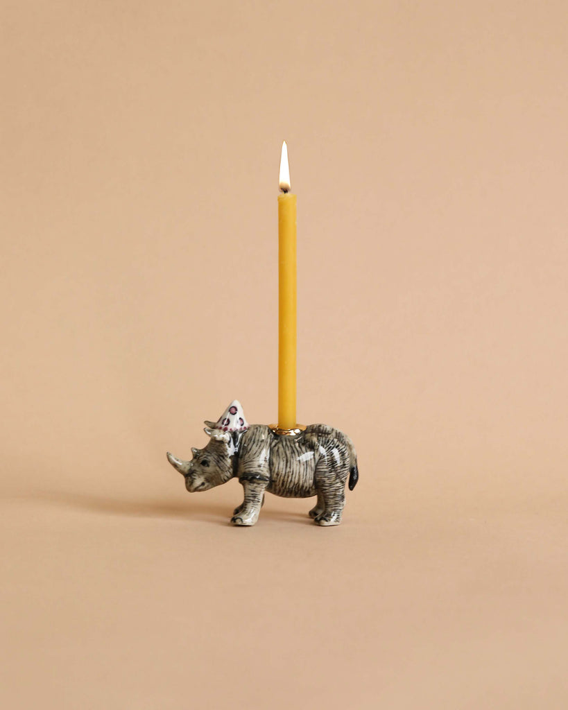 A unique Rhino Cake Topper shaped like a rhinoceros, hand-painted with detailed stripes and a pink floral pattern on its back, supporting a lit birthday candle. The background is plain, light.