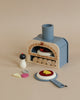 A children's Wooden Make Me A Pizza Set, including a pizza oven, some detachable burners, and wooden play food like a pizza slice with various toppings and an egg, on a soft beige background.