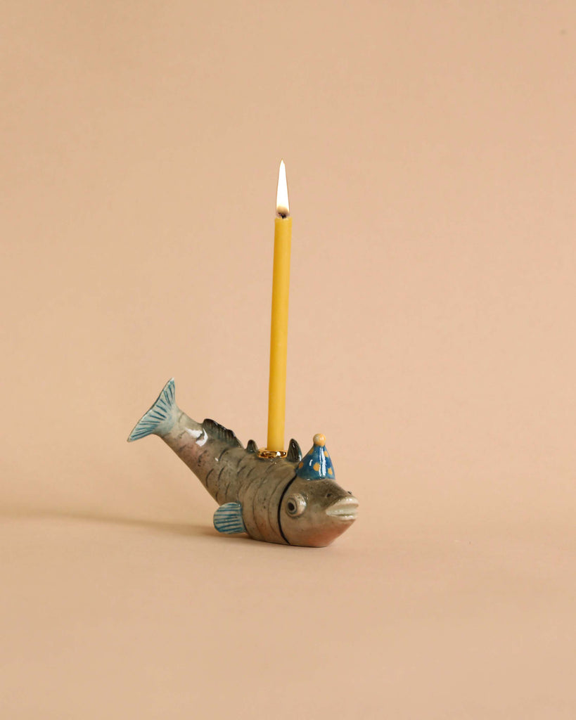 A whimsical hand-painted porcelain Fish Cake Topper shaped like a fish, with a yellow pencil inserted into it, positioned against a plain beige background.