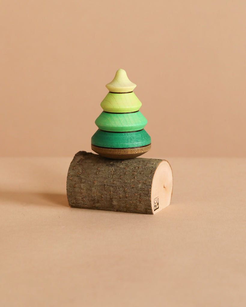 The Mader Spinning Tree Top On Trunk, a small green pine tree toy crafted from Maple wood, is stacked on a short cylindrical piece of tree branch against a plain beige background.