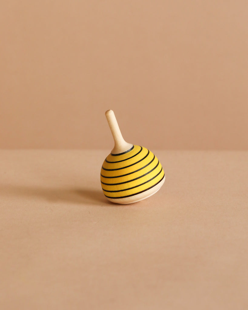 The Spinning Top - Bumble Bee, crafted from maple wood with yellow and black stripes, sits tilted on a plain beige surface against a matching beige background.