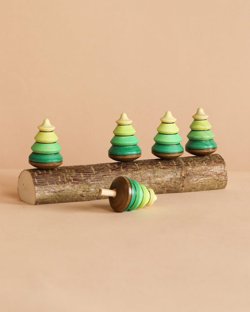 Four small green maple wood Mader Spinning Tree Top Forest On Trunk toys stand on a log, with one more tree lying in front, all displayed on a beige background.