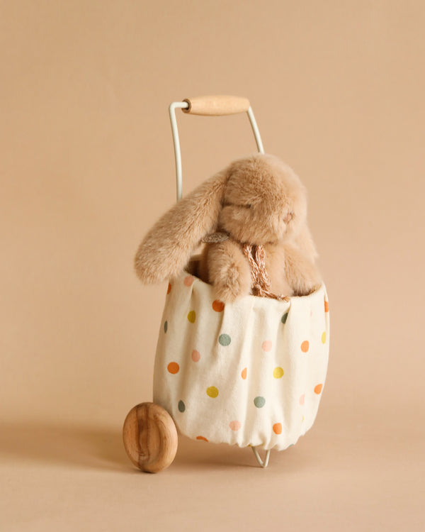 A Maileg Mini Bunny & Trolley Set peeking out from a polka-dotted washable fabric basket with a wooden wheel, set against a soft beige background.