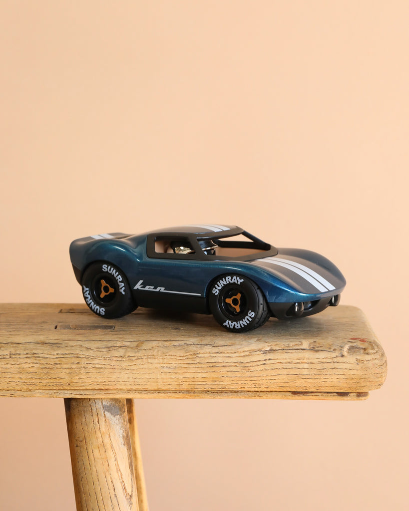 The Playforever MINI KEN CASH Blue racing car with white stripes rests on a wooden stool against a beige background. This classic toy features "SUNRAY" on its tires, inspired by Ken Miles' legendary race cars.