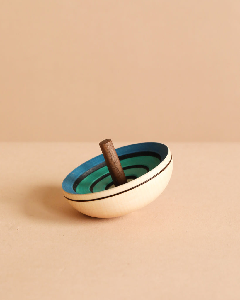 The Mader UFO Flora Spinning Top - Blue, crafted from maple wood with a dark handle and ombre green and blue concentric circles, rests on a beige surface against a coordinating beige background.