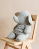 A soft gray Maileg Big Grey Elephant plush toy sitting on a wooden chair against a light beige background. The toy is facing away, showing its back and large floppy ears.