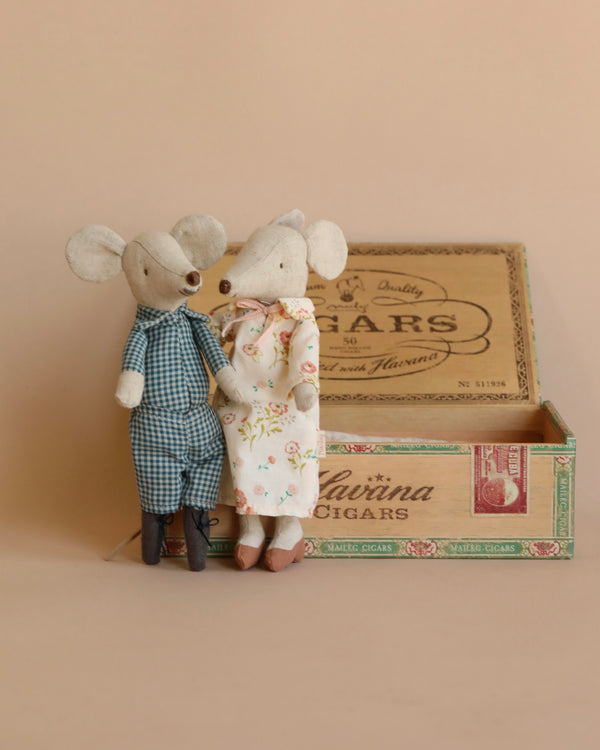 Two Maileg Grandma & Grandpa Mice in Cigarbox, dressed in human clothes, are standing and embracing each other beside an open vintage cigar box. The background is a plain, light peach color.