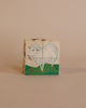 A Wooden Block Puzzle - 4 Piece Domestic Animals featuring a picture of a sheep standing in a grassy field, with parts of images on other sides visible at the edges. The background is a soft beige.