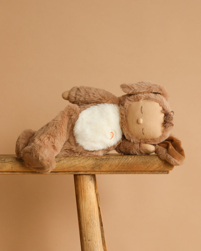 The Olli Ella Cozy Dinkum - Dog Digby, a plush toy with brown fur, floppy ears, and a white belly, lies on its side atop a rustic wooden bench set against a beige background.