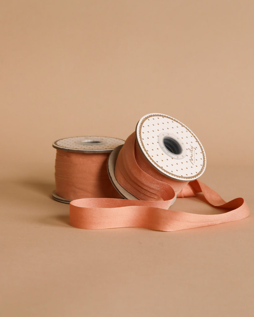 Two rolls of Maileg Ribbon - Peach Rose (10m), one upright and one partially unspooled, stand out against a beige background—ideal for elegant gift wrapping or crafts.