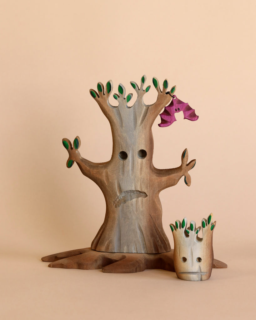 The Handmade Wooden Haunted Pumpkin Cottage Set features a wooden tree with green leaves and a sad face beside a smaller neutral tree stump. A purple bird perches on a branch—all perfect for Montessori imaginative play against a beige background.