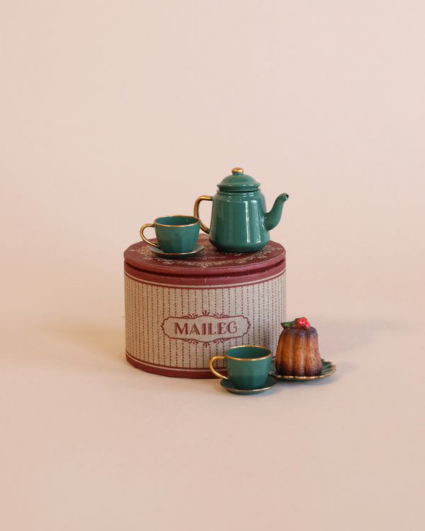 A Maileg Christmas Tea Party with a green metal teapot, two matching teacups, a cake, and saucers is arranged on a vintage round tin container labeled "Maileg." Perfect for the mouse family size, the background is a plain, light beige color.