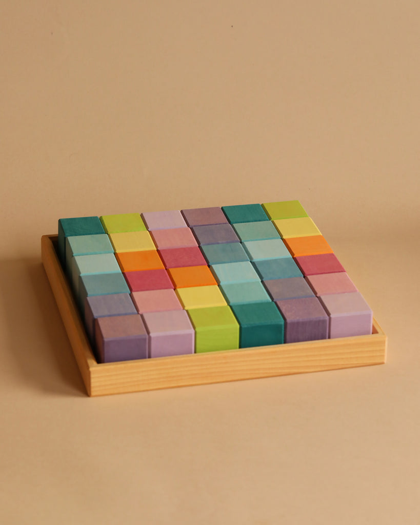 A wooden tray filled with a grid of colorful Grimm's Pastel Mosaic Building Blocks arranged neatly. Perfect for children, these blocks come in various shades of green, blue, purple, pink, orange, and yellow. They create a vibrant and orderly pattern against a plain beige background while enhancing fine motor skills.