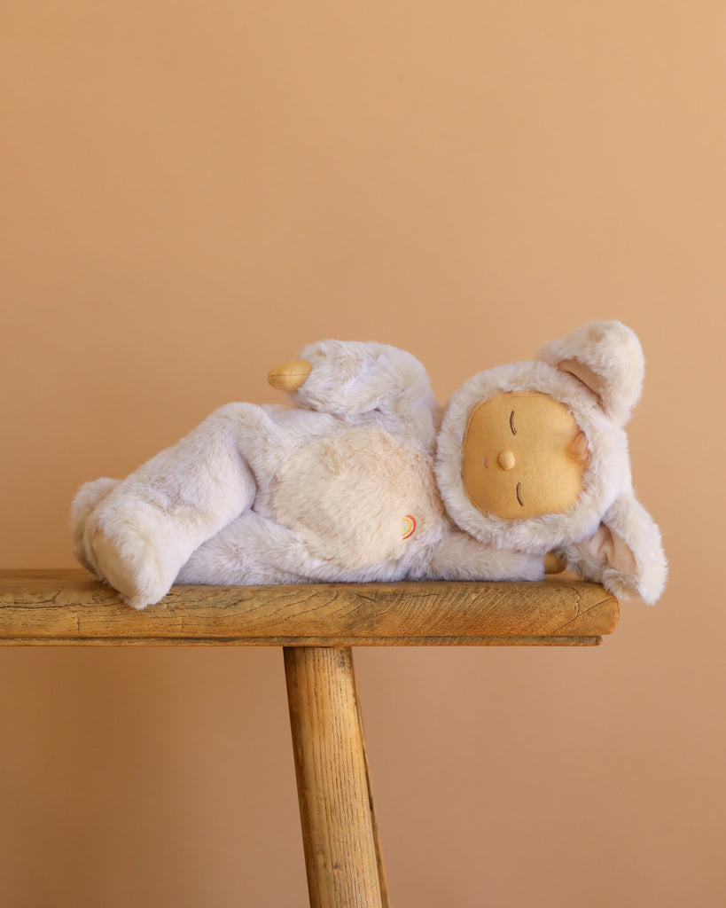 The Olli Ella Cozy Dinkums Fox - Fenix plush, dressed in a soft light purple animal outfit, rests on its side on a wooden stool with a plain beige backdrop.