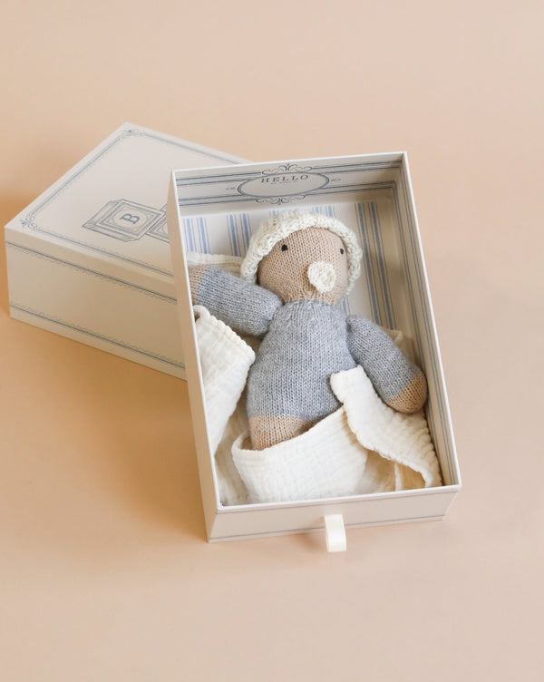 A handcrafted Hand-Knit Baby Doll - Beige in blues and whites, placed carefully inside an elegant keepsake box against a soft peach background.