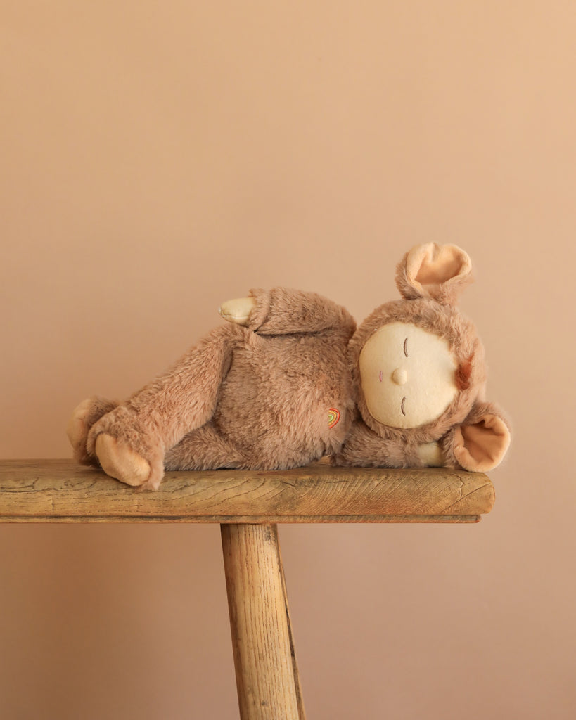 The Olli Ella Cozy Dinkum - Mousy Pickle plush toy, featuring soft brown fur and bunny ears, lies peacefully on a wooden bench with its eyes closed, set against a plain peach background.