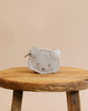 A small Donsje Mini Leather Backpack - Cat designed to look like a cat's face, with stitched details and pointy ears, resting on a rustic wooden stool against a light beige background.